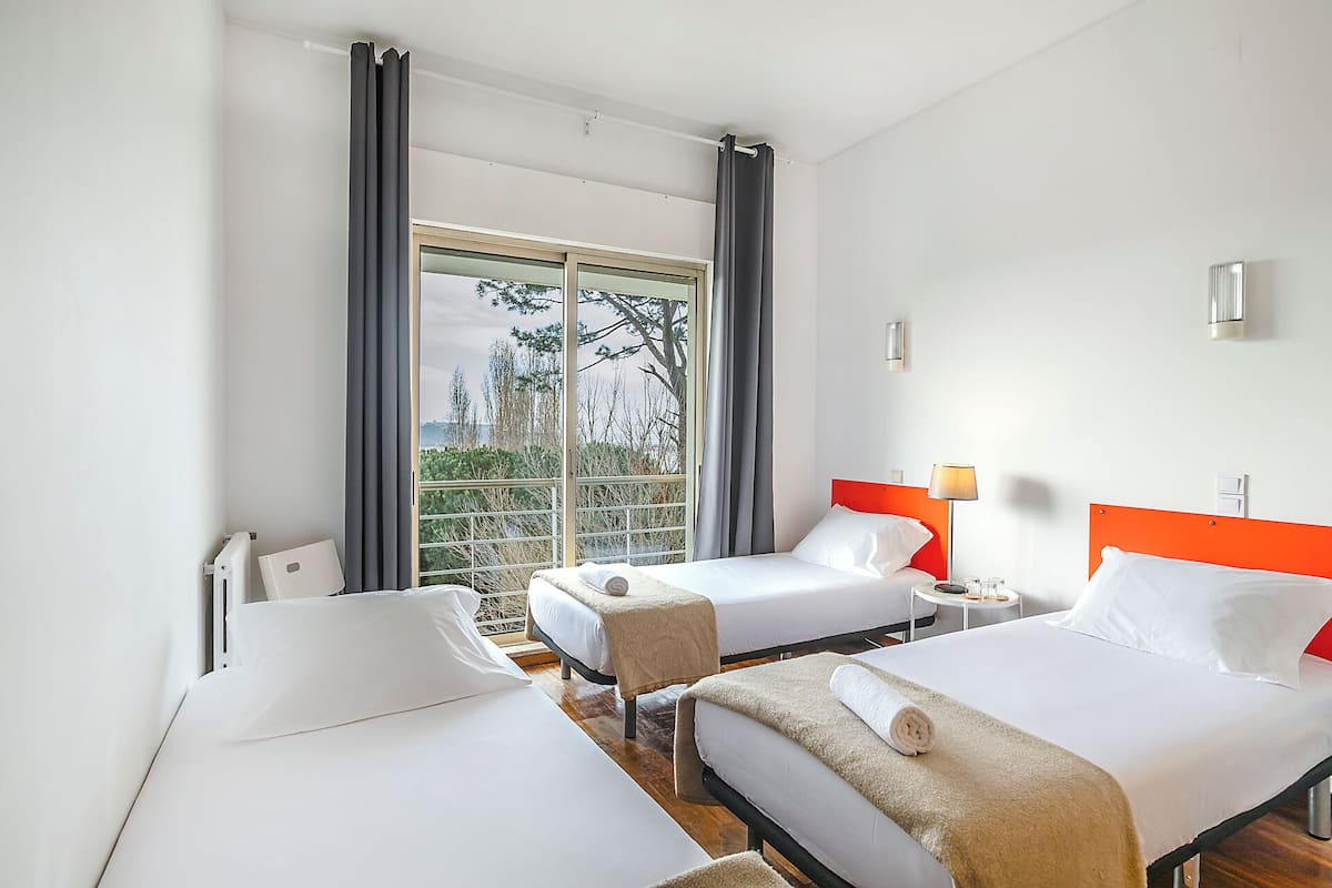 Une chambre d’hôtel avec deux lits, une fenêtre avec des rideaux, une vue sur les arbres et une petite table avec une lampe.