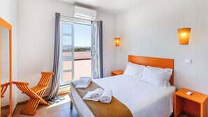 Une chambre d’hôtel comprenant un lit, une chaise, une table de chevet, un appareil de climatisation et une vue sur l’extérieur, visible à travers une porte vitrée.
