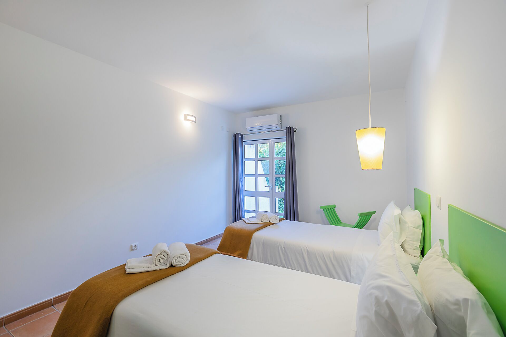 A hotel room with two beds, a window with curtains, a green headboard, and a wall-mounted air conditioner.