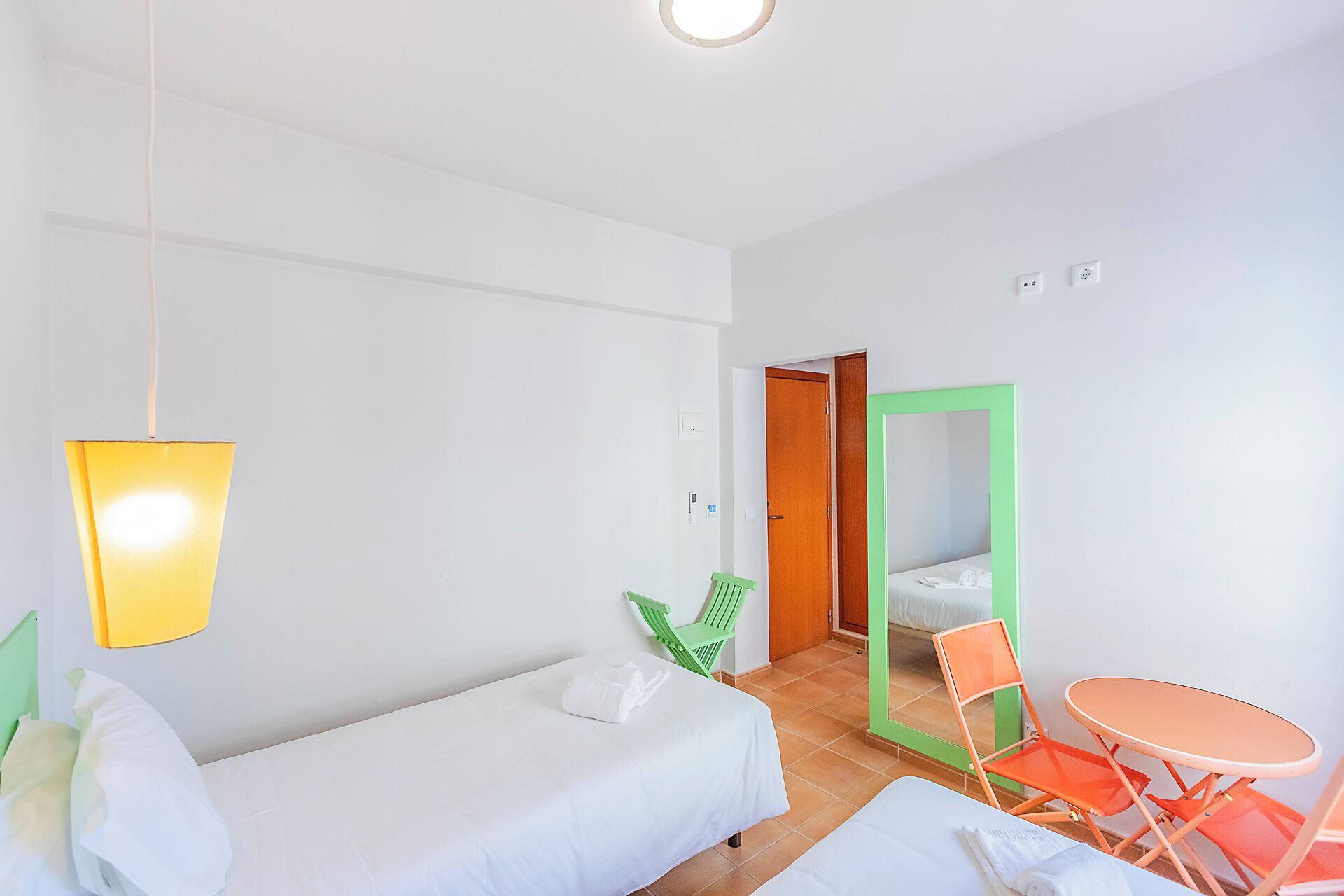 A hotel room with a bed, a small table, two chairs, a hanging light fixture, and a door leading to another room.