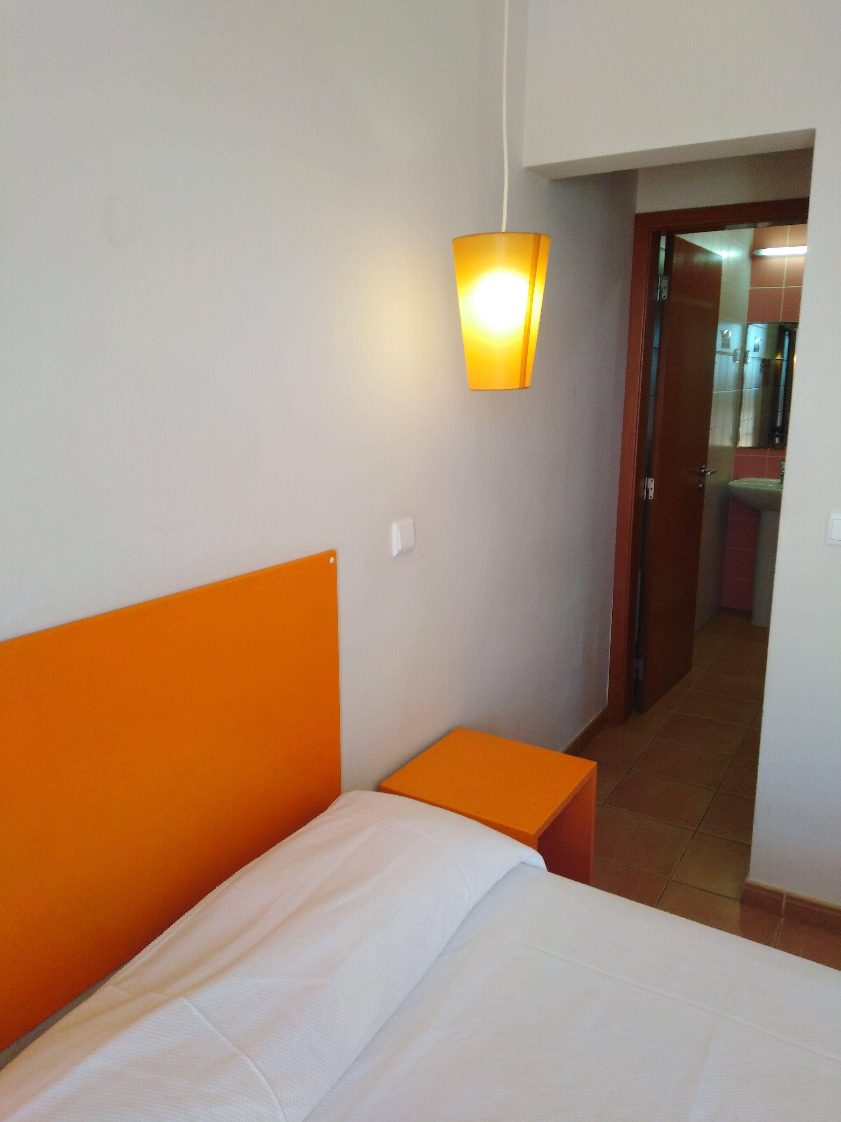 Une chambre d’hôtel avec un lit, une table de chevet orange, une lampe fixée au mur et une vue sur la salle de bain par une porte ouverte.