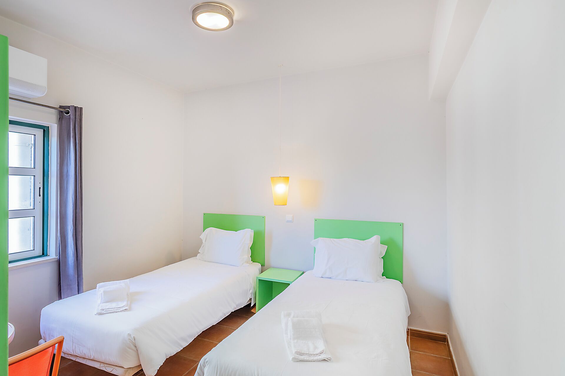 A hotel room with two single beds, a small table, a chair, a window with curtains, and a wall-mounted light fixture.