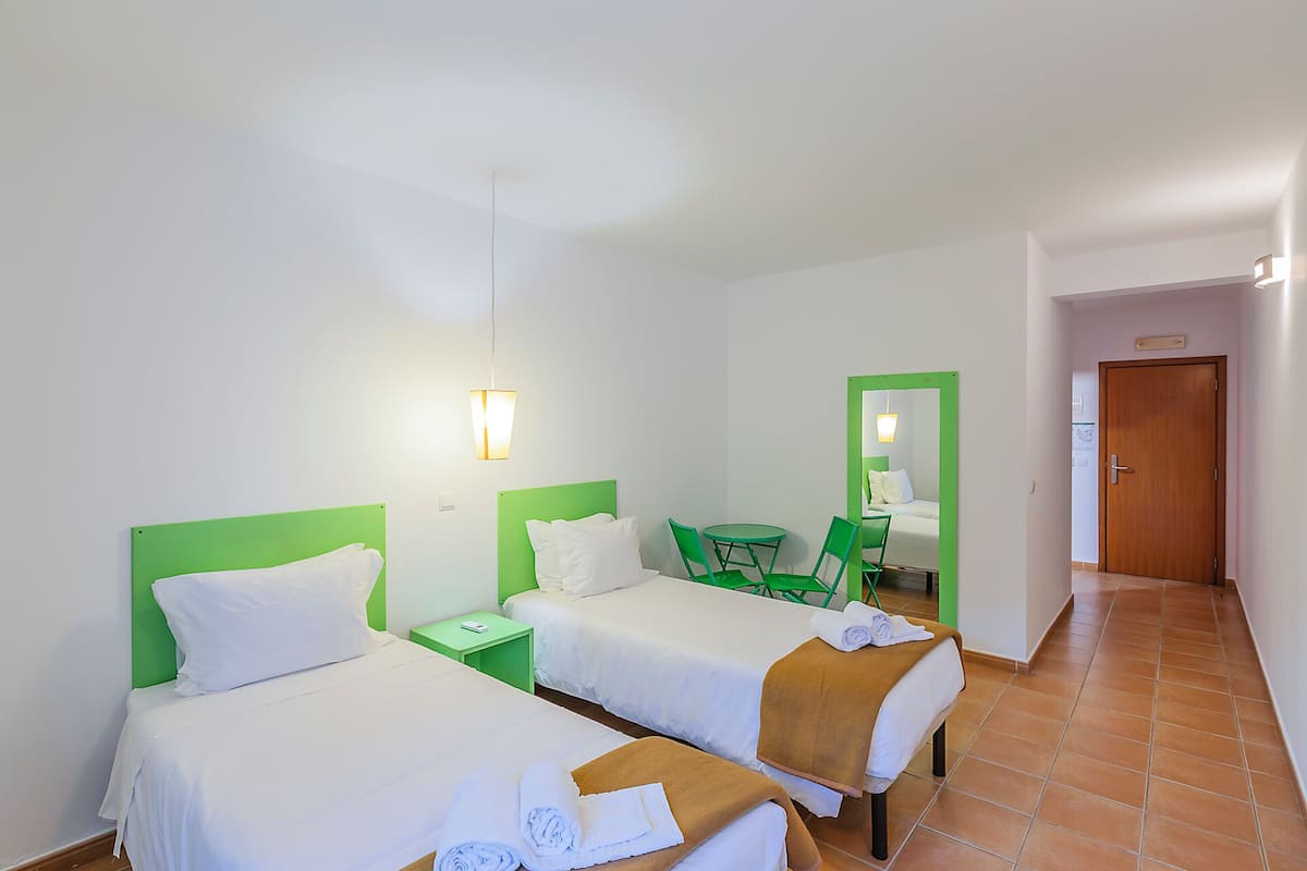 Habitación de hotel con dos camas, un espejo de marco verde, una mesita con sillas y una puerta de madera.