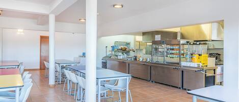 A modern kitchen with stainless steel appliances, a dining area with white chairs and tables, and a tiled floor.
