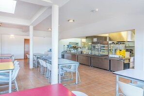 Una cocina moderna con electrodomésticos de acero inoxidable, un comedor con sillas y mesas blancas, y un suelo de baldosas.