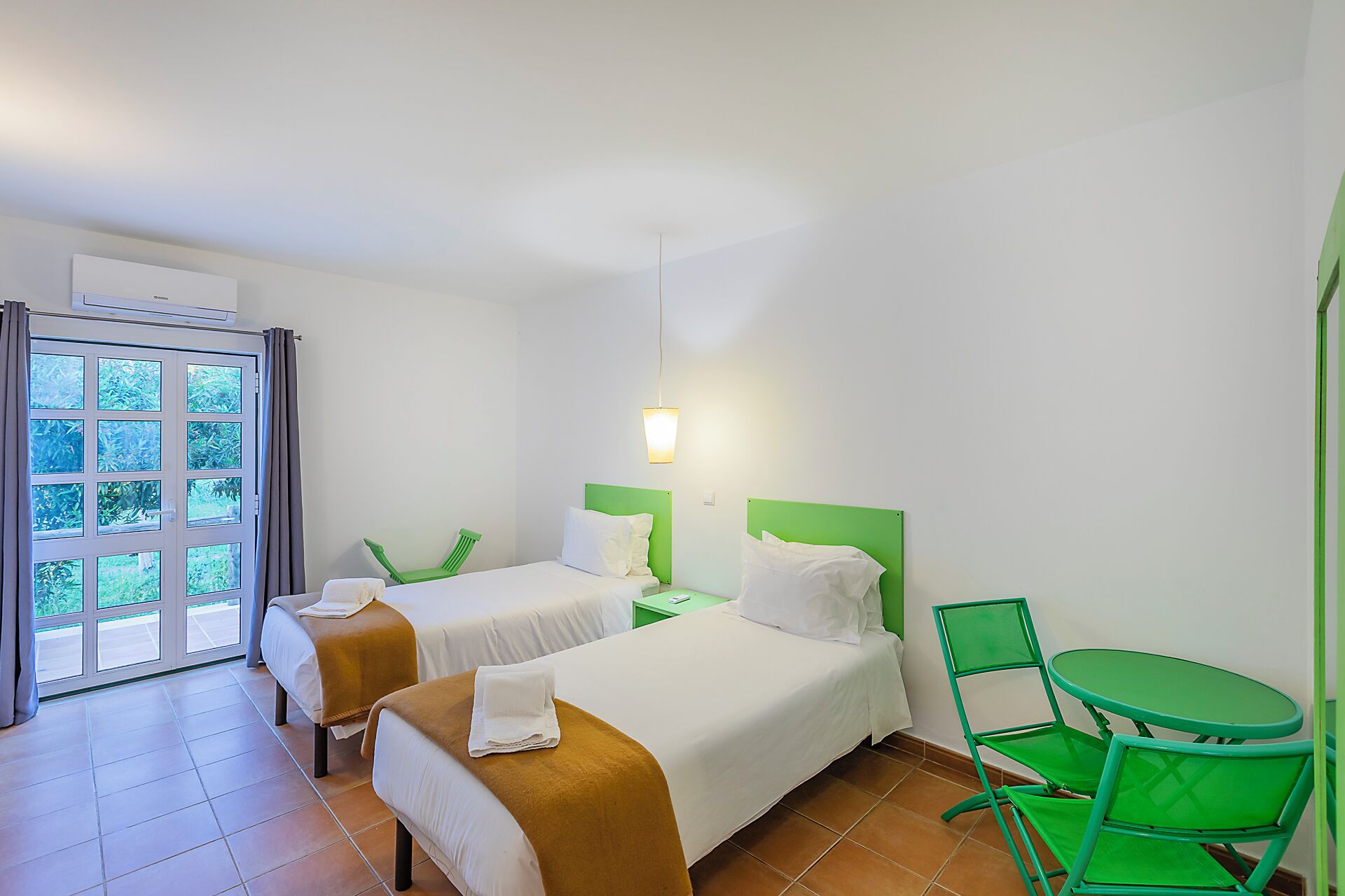 Une chambre d’hôtel avec deux lits, un fauteuil vert, une petite table et une fenêtre avec des rideaux.