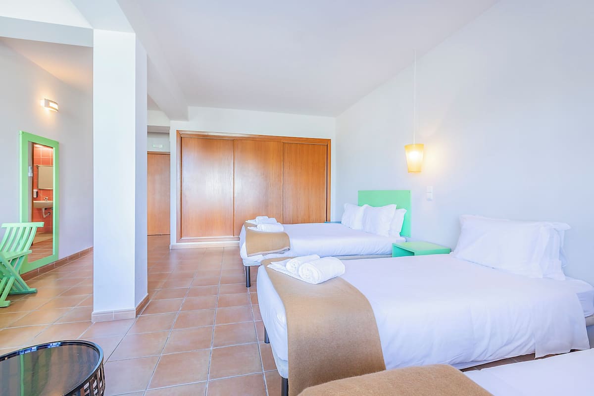 Habitación de hotel con tres camas, una silla verde, una mesita pequeña y un armario de madera.