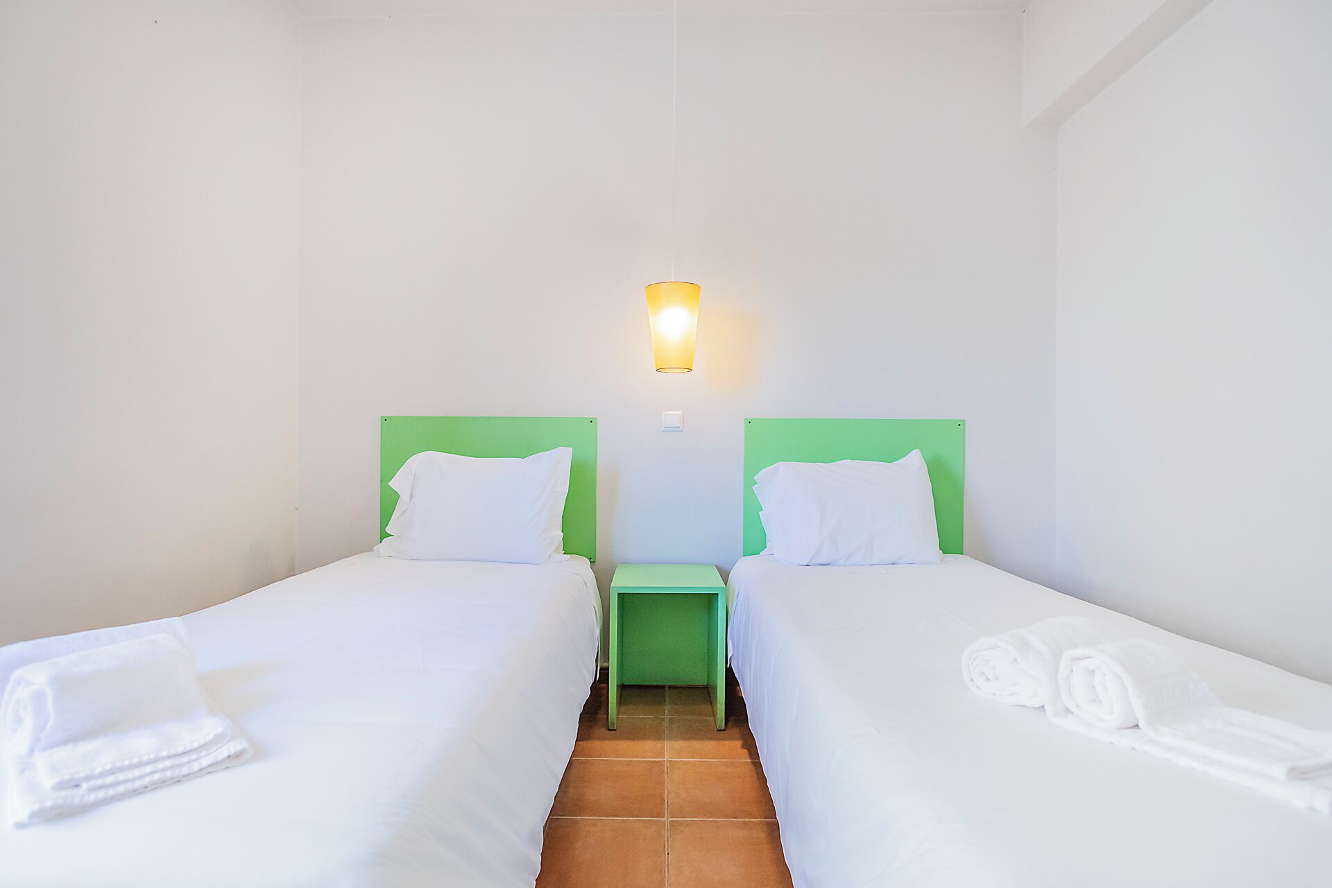 A twin room with two beds, a nightstand, a wall-mounted light, and a rolled towel on each bed.