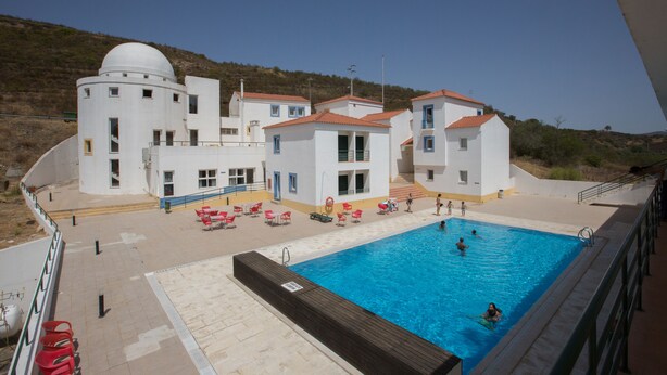 A swimming pool area with a large pool, adjacent buildings, and a hillside in the background. - HI Alcoutim – Pousada de Juventude - Hostel (Alcoutim)