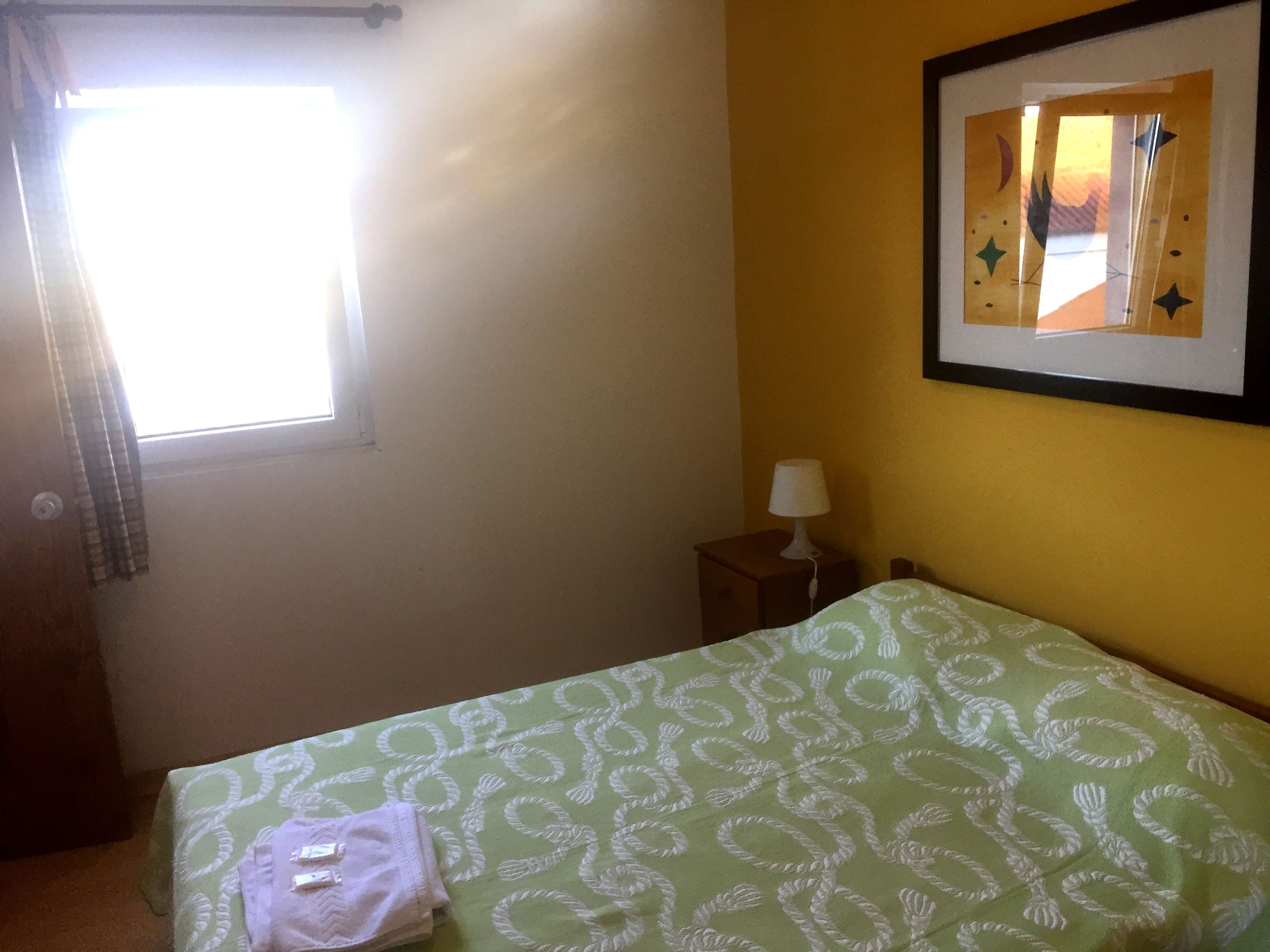 A room with a bed, a nightstand, a lamp, a window with curtains, and a framed picture on the wall.
