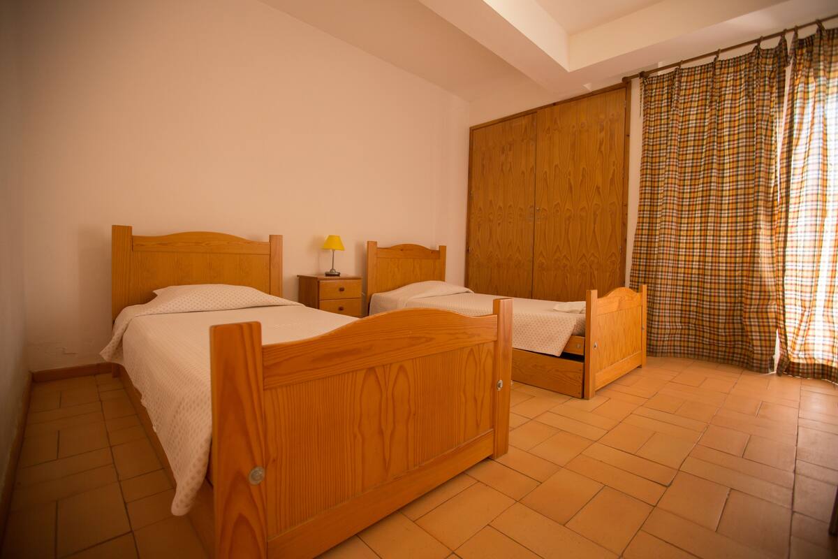 A bedroom with two wooden beds, a wardrobe, and a window with checkered curtains.