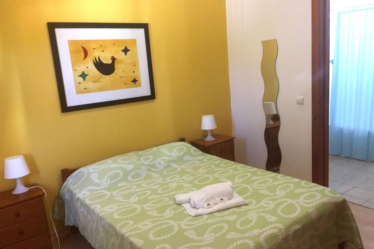A room with a bed, two bedside tables, a framed picture on the wall, and a door leading to another room.