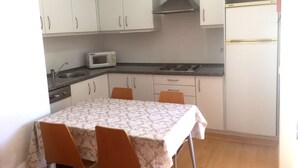 A compact kitchen with white cabinets, a microwave, a stove, and a refrigerator. There is a table with a patterned tablecloth and two chairs. - HI Alcoutim – Pousada de Juventude - Hostel (Alcoutim)