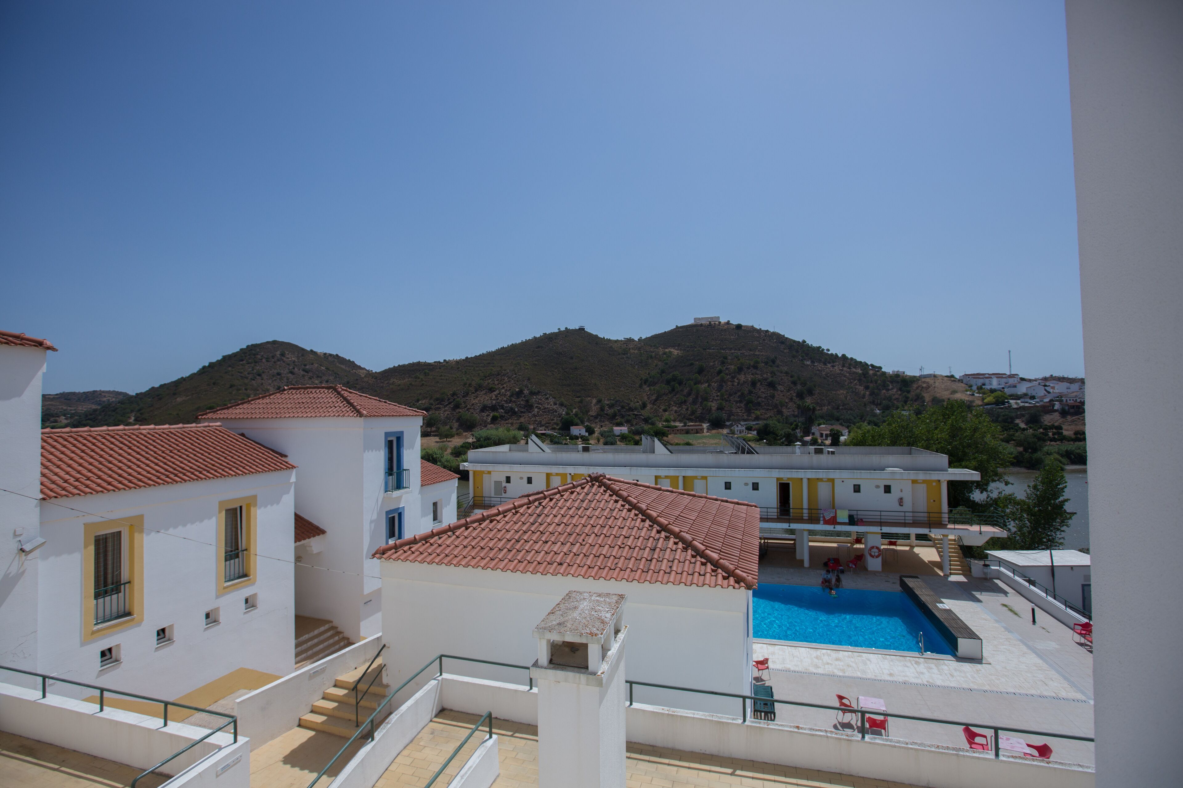 A residential area with a swimming pool, white buildings with red roofs, and a hilly landscape in the background.