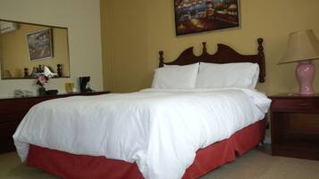 Una cama bien hecha con ropa de cama blanca y falda roja, un cabecero de madera, una mesita de noche con una lámpara, un espejo y un cuadro de una escena costera en la pared.