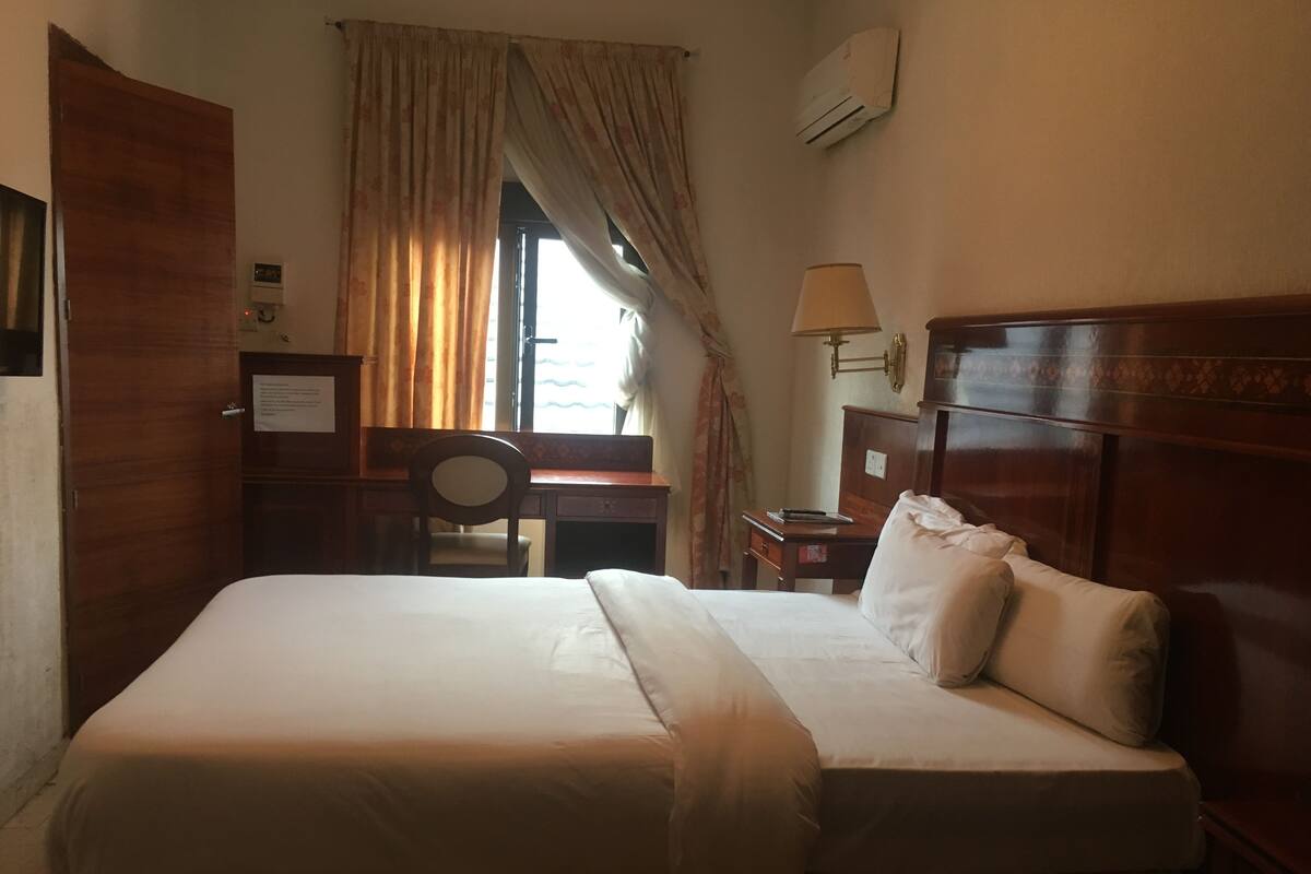 A neatly made bed with a wooden headboard, a desk with a chair, a lamp, and an air conditioning unit on the wall.