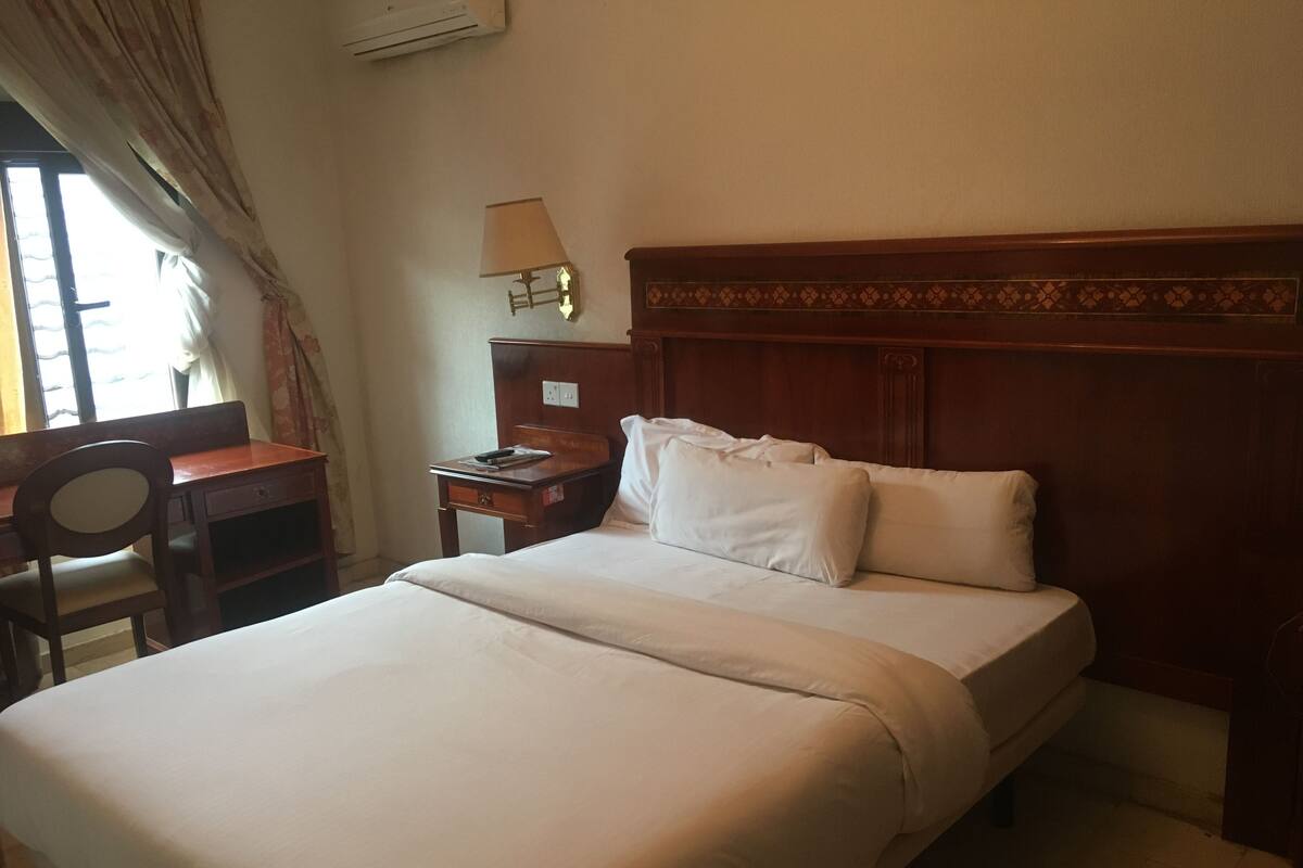 A hotel room with a bed, a desk, a chair, a nightstand, and a lamp.