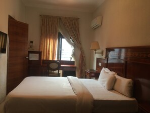 A hotel room with a bed, desk, chair, lamp, and window with curtains. - Hotel De Kajola (Lekki)