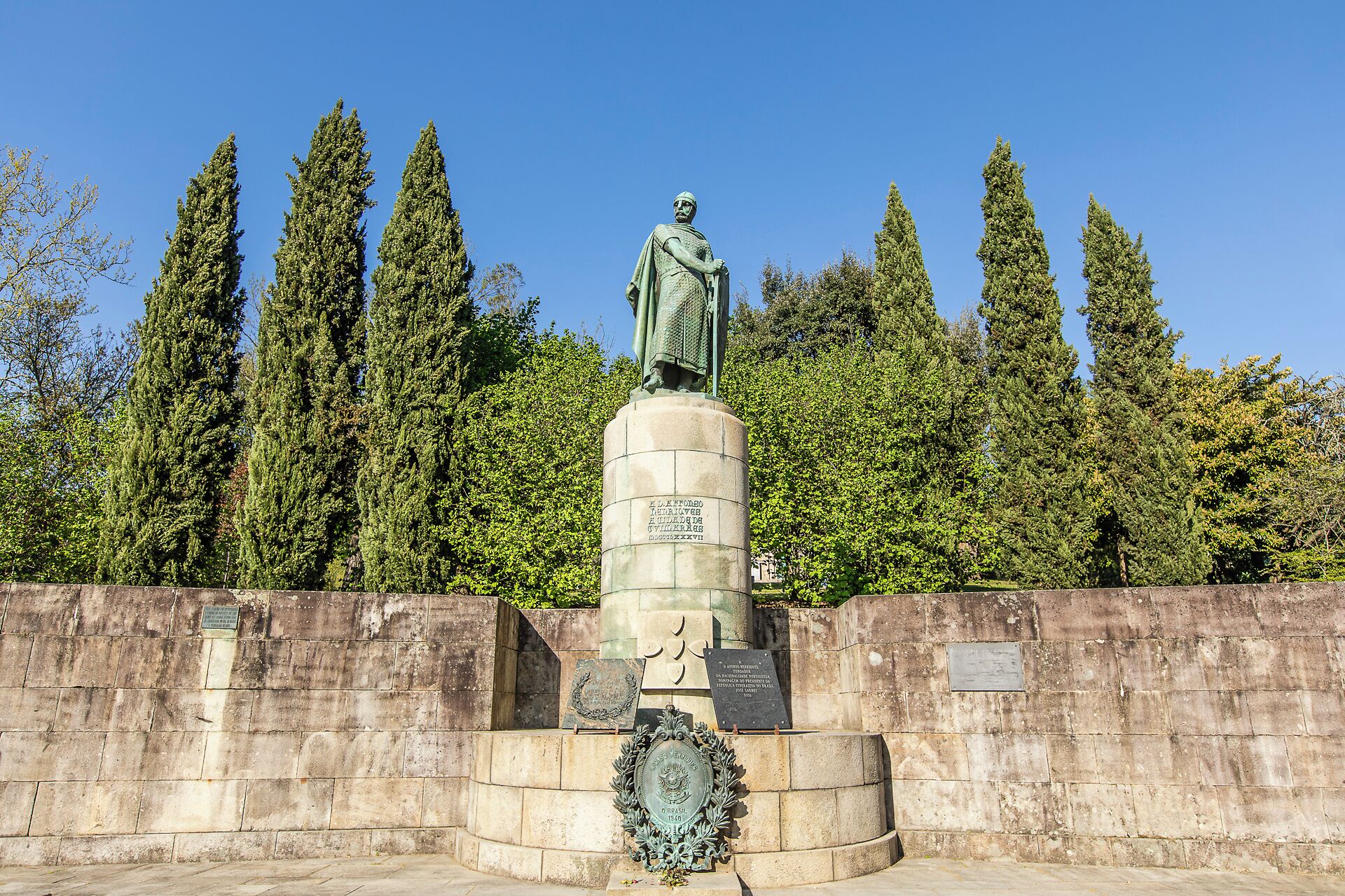 Eine Statue auf einem Sockel mit Inschriften, umgeben von einer Steinmauer und hohen Bäumen.