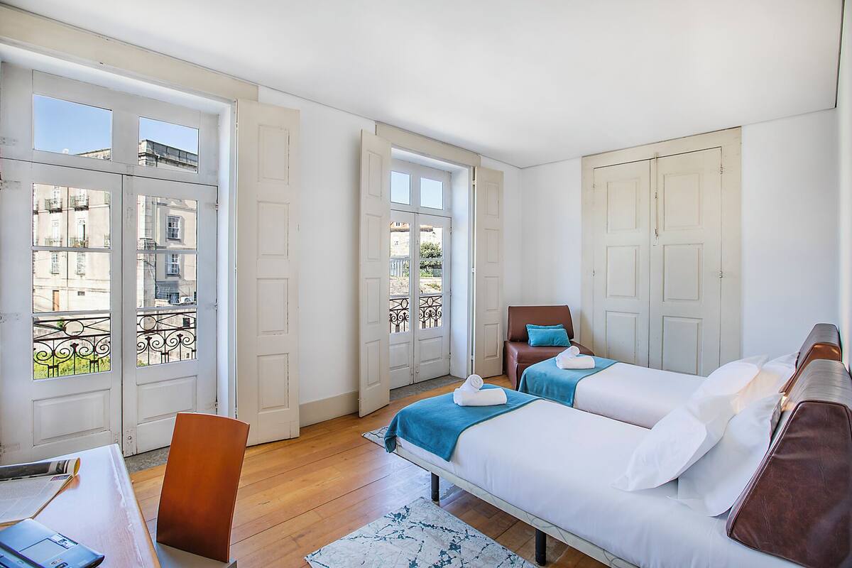 Una habitación con dos camas, piso de madera, un escritorio con silla y ventanas grandes.