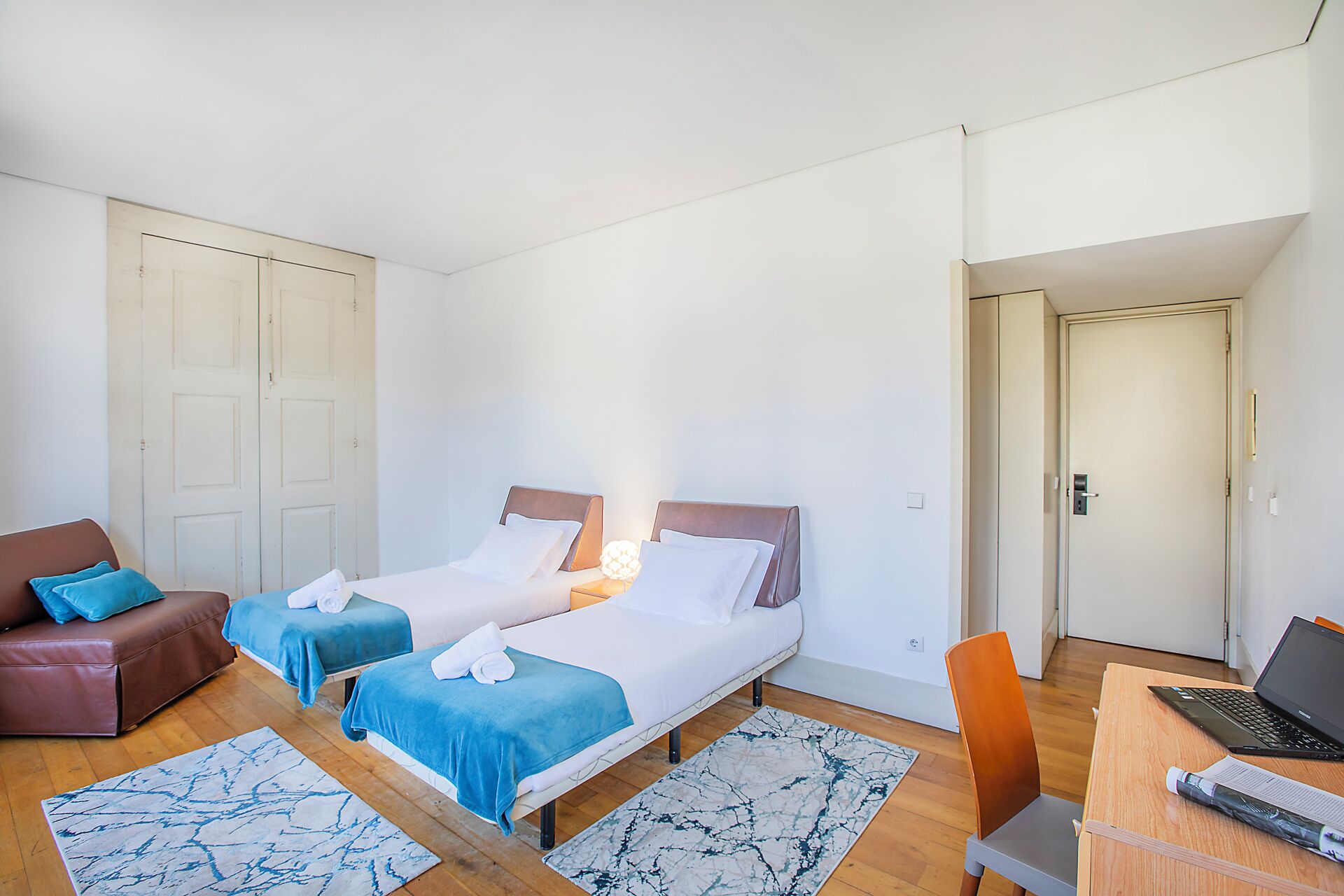 A hotel room with two beds, a sofa, a desk with a laptop, and a door leading to another room.