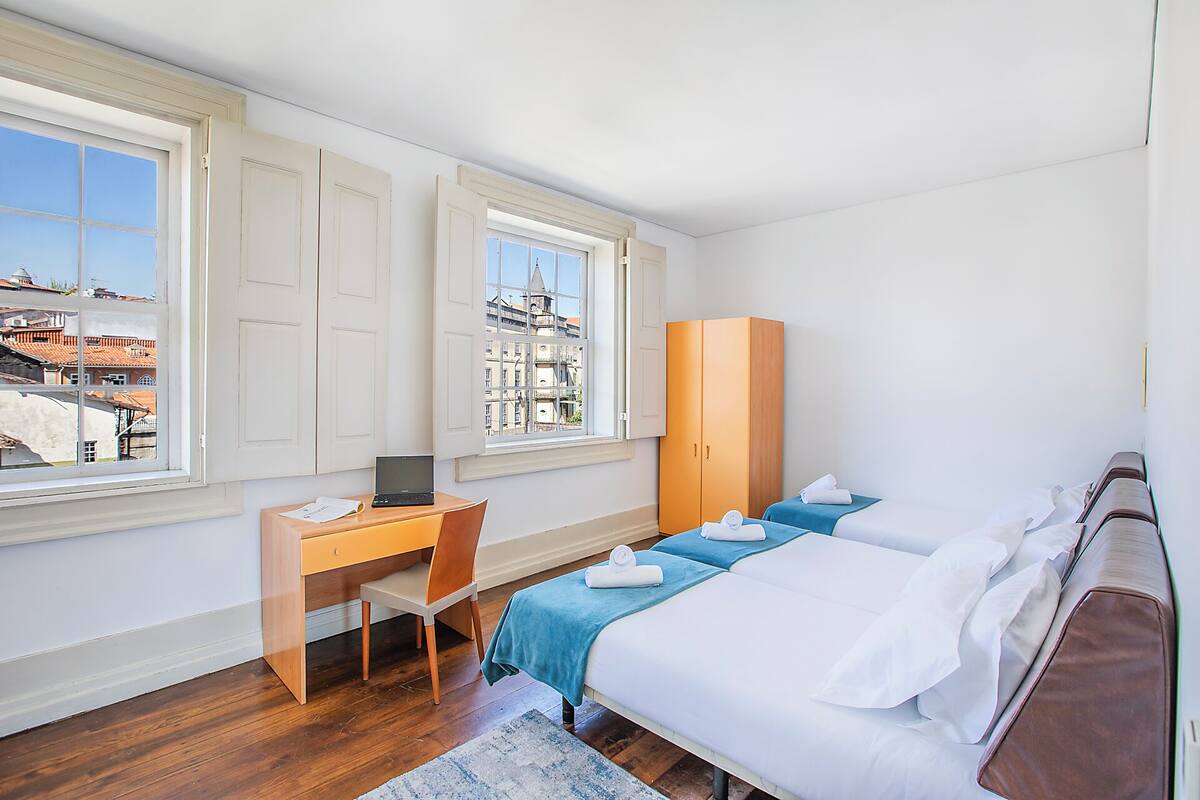Habitación de hotel con escritorio, dos camas, armario y vistas a edificios a través de las ventanas.