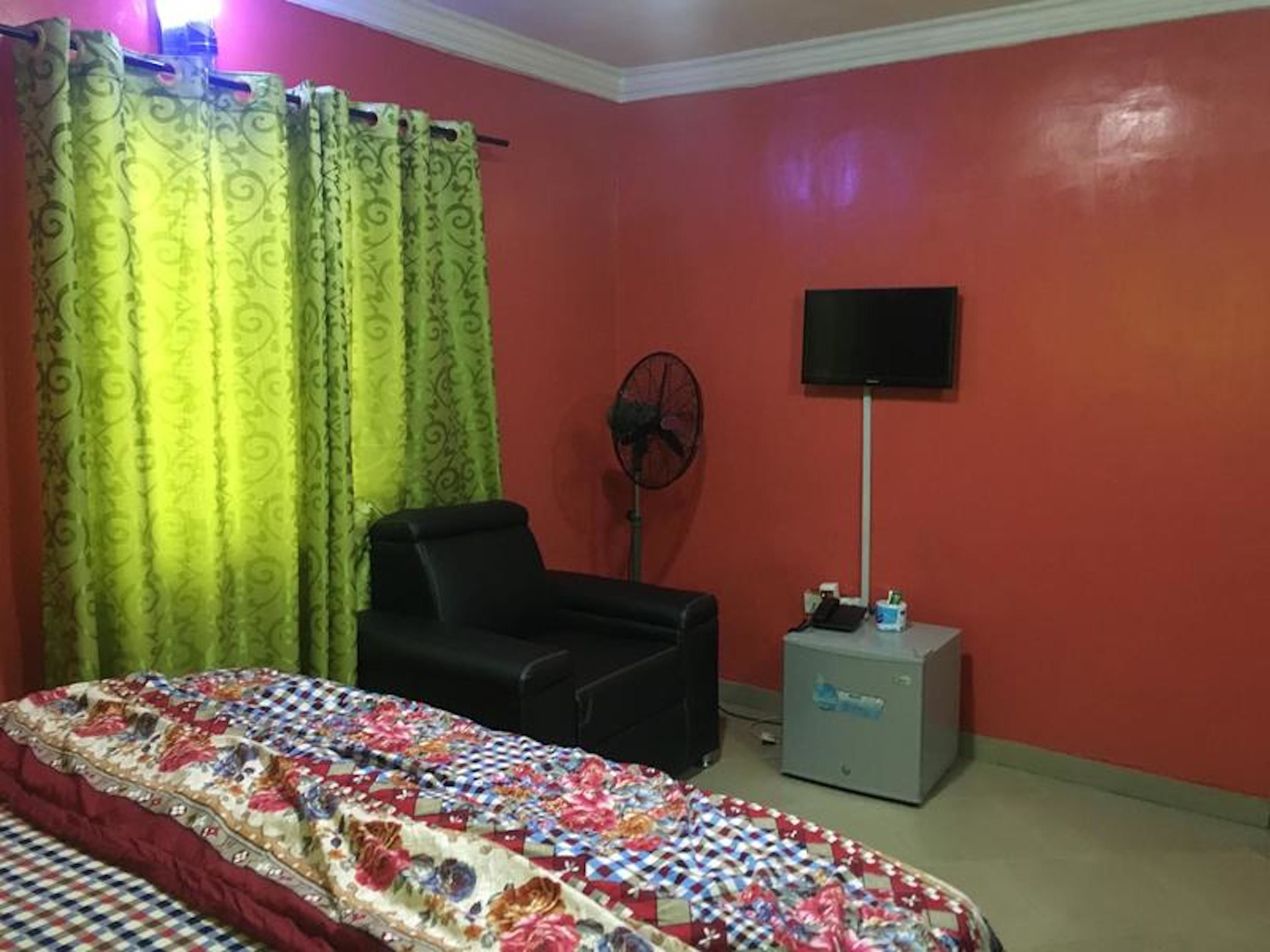 A room with a bed, a black chair, a standing fan, a television on the wall, and a small cabinet.