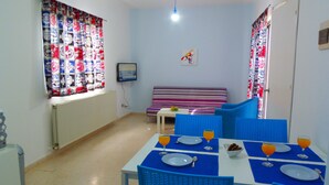 Una habitación pequeña y luminosa con una mesa de comedor preparada para dos personas, un sofá y un televisor en la pared.