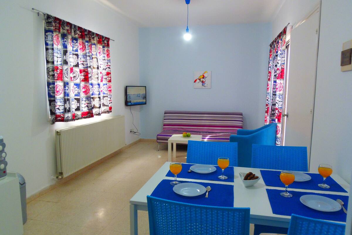 Una habitación pequeña y luminosa con una mesa de comedor preparada para dos personas, un sofá y un televisor en la pared.