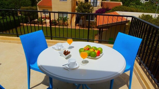 Una mesa redonda blanca con sillas azules en un balcón, un plato de fruta, un vaso de zumo de naranja y un pequeño bol de frutos secos.