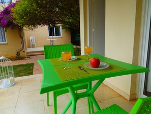 A green outdoor dining area with a table set for two, featuring a glass of orange juice and an apple on a plate.