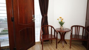 A room with a wooden door, two wooden chairs, a small round table with a vase of yellow flowers, and a view of a cityscape through a window with curtains. - Orange Quy Nhon (Quy Nhon Nam)