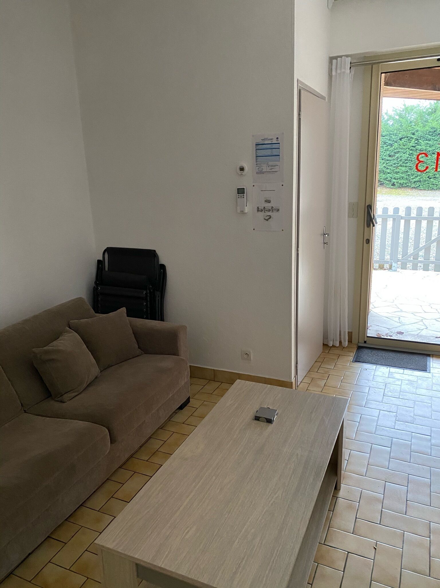 Una habitación con un sofá color crema, una mesa de centro de madera, una puerta con una cortina blanca y vistas a una valla blanca en el exterior.