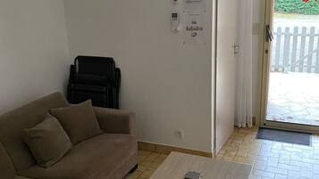Una habitación con un sofá color crema, una mesa de centro de madera, una puerta con una cortina blanca y vistas a una valla blanca en el exterior.