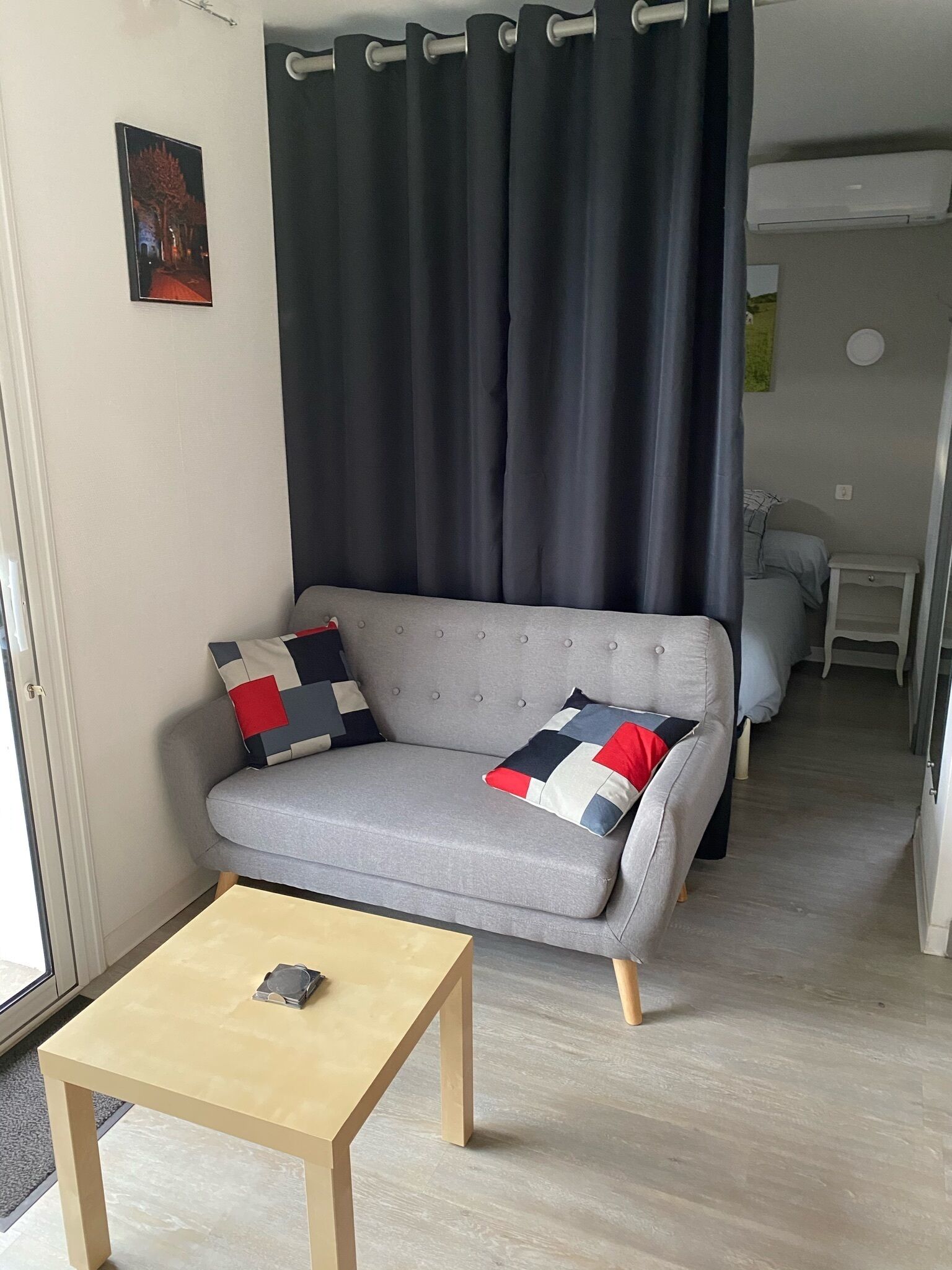 Ein Wohnzimmer mit grauem Sofa, zwei gemusterten Kissen, einem Holztisch und einem dunklen Vorhang.