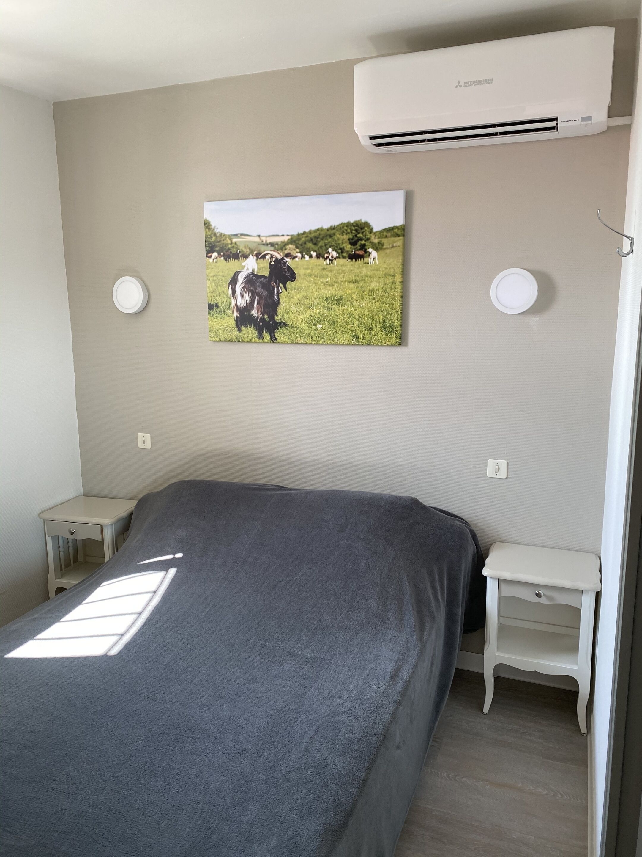 Ein Schlafzimmer mit Bett, Nachttisch, Klimaanlage und einem Wandbild, das eine Kuh auf einer Wiese zeigt.