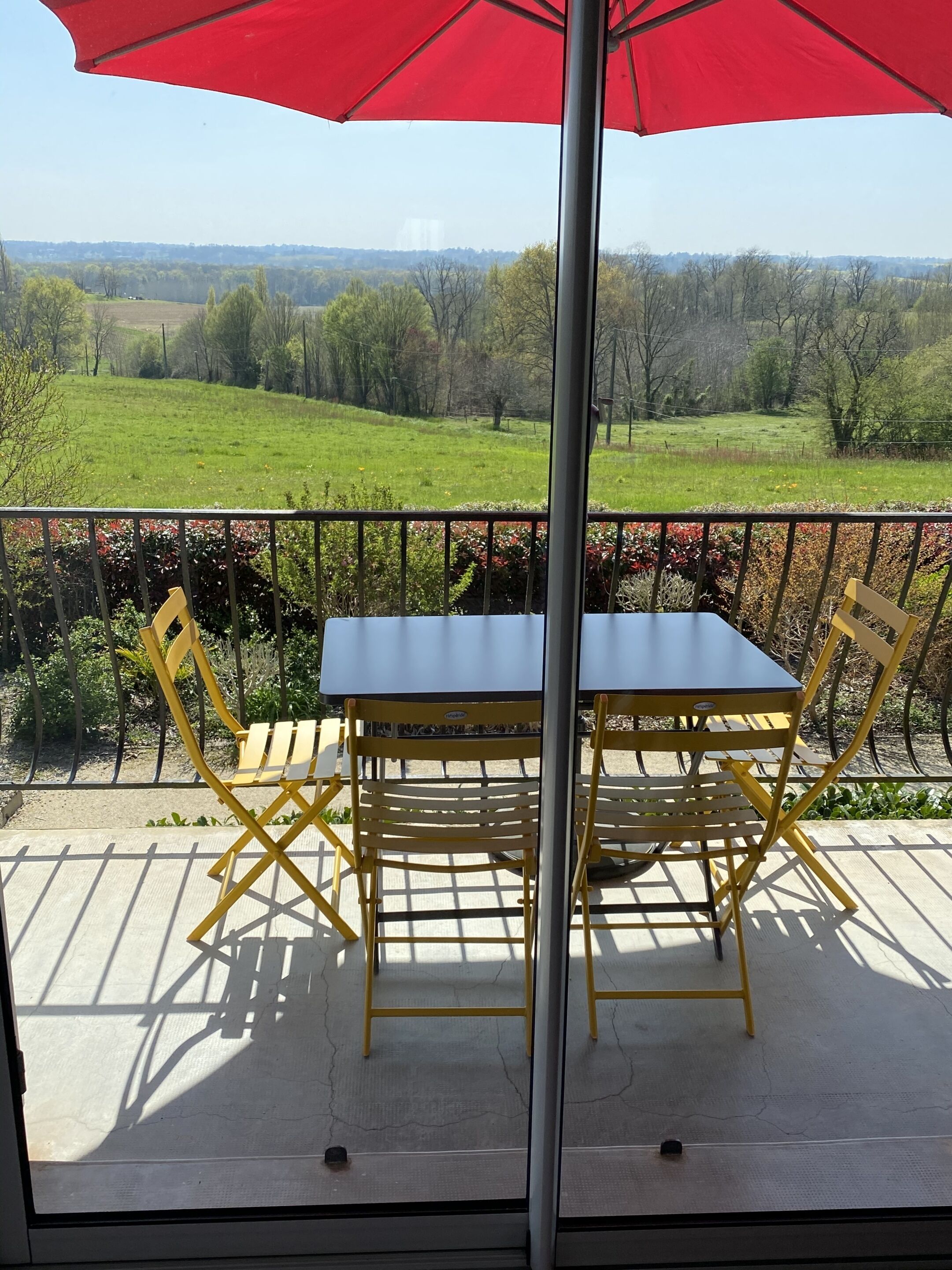 Un balcón con mesa y sillas, una sombrilla roja y vistas a un campo verde y árboles.