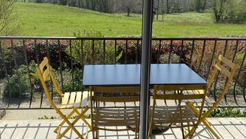 Un balcón con mesa y sillas, una sombrilla roja y vistas a un campo verde y árboles.