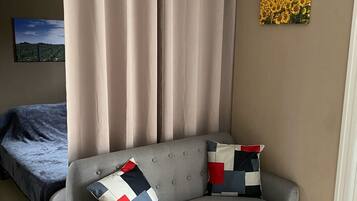 Una habitación con un sofá gris, dos almohadas estampadas, una mesa de madera, una cama con cubrecama azul, cortinas color beige y un cuadro colgado en la pared con girasoles.