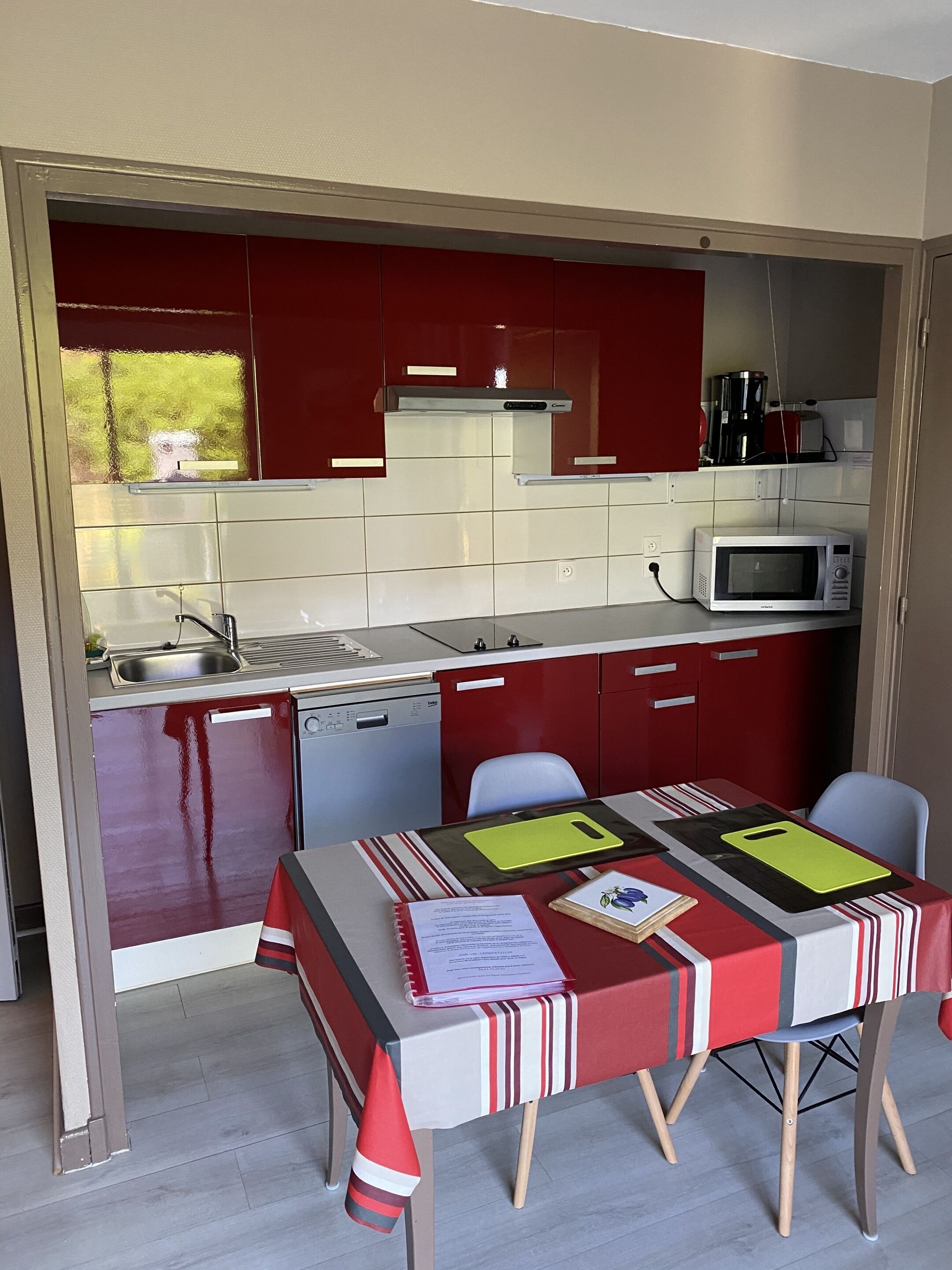 A compact kitchen with red cabinets, a microwave, and a sink. There is a dining table with a striped tablecloth and two chairs.
