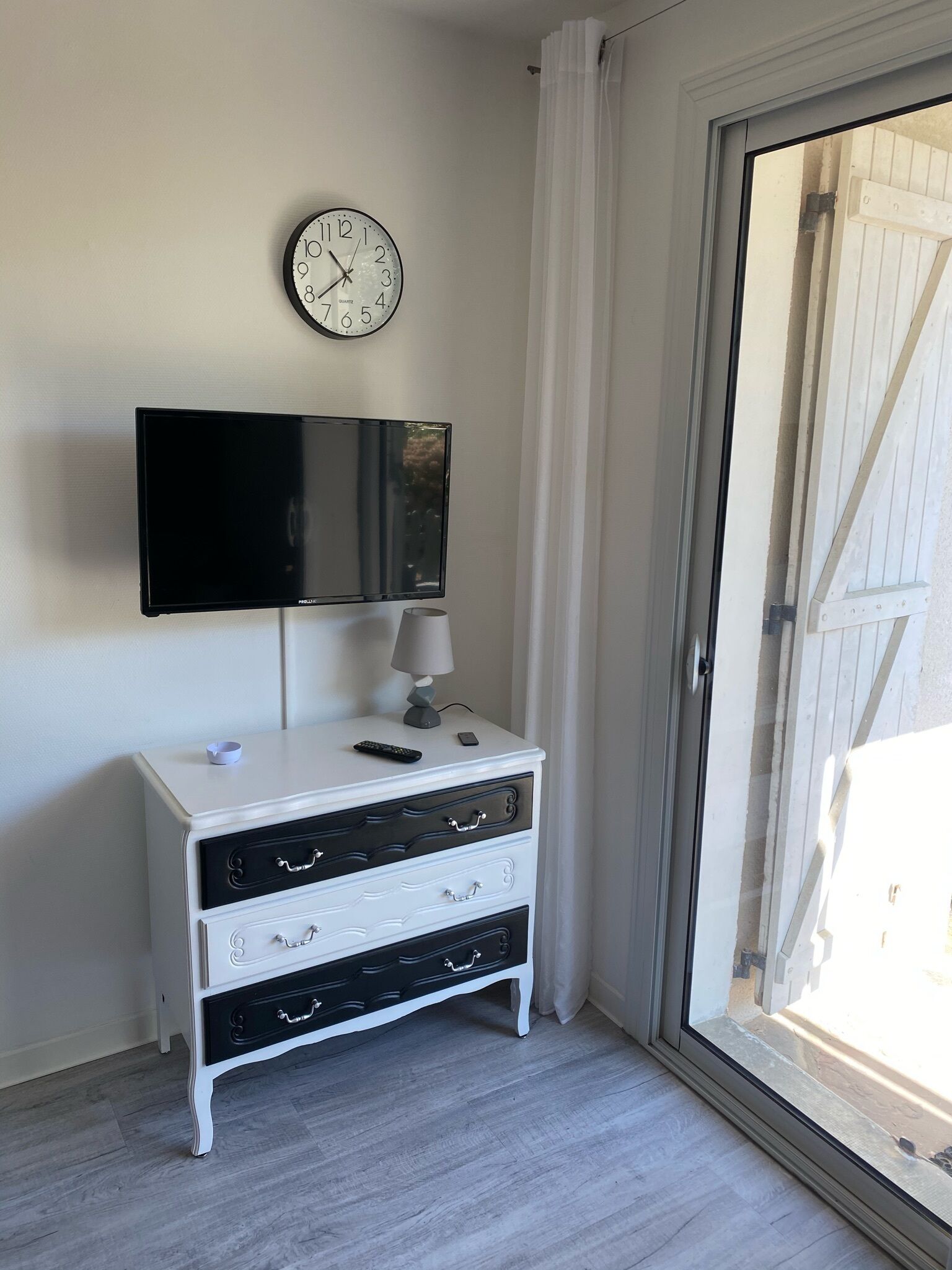 Una habitación con un aparador blanco, un televisor de pantalla plana en la pared, una lámpara sobre el aparador y un ventanal corredero que da a un espacio exterior.