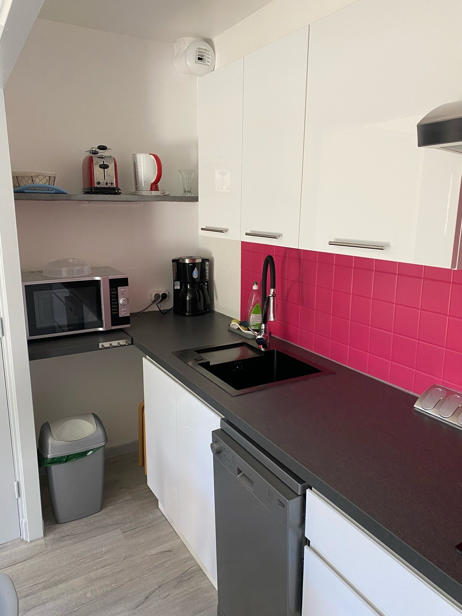 Une cuisine compacte avec des meubles blancs, un plan de travail noir, un four à micro-ondes, une machine à café et un évier avec un pare-éclaboussures rose.