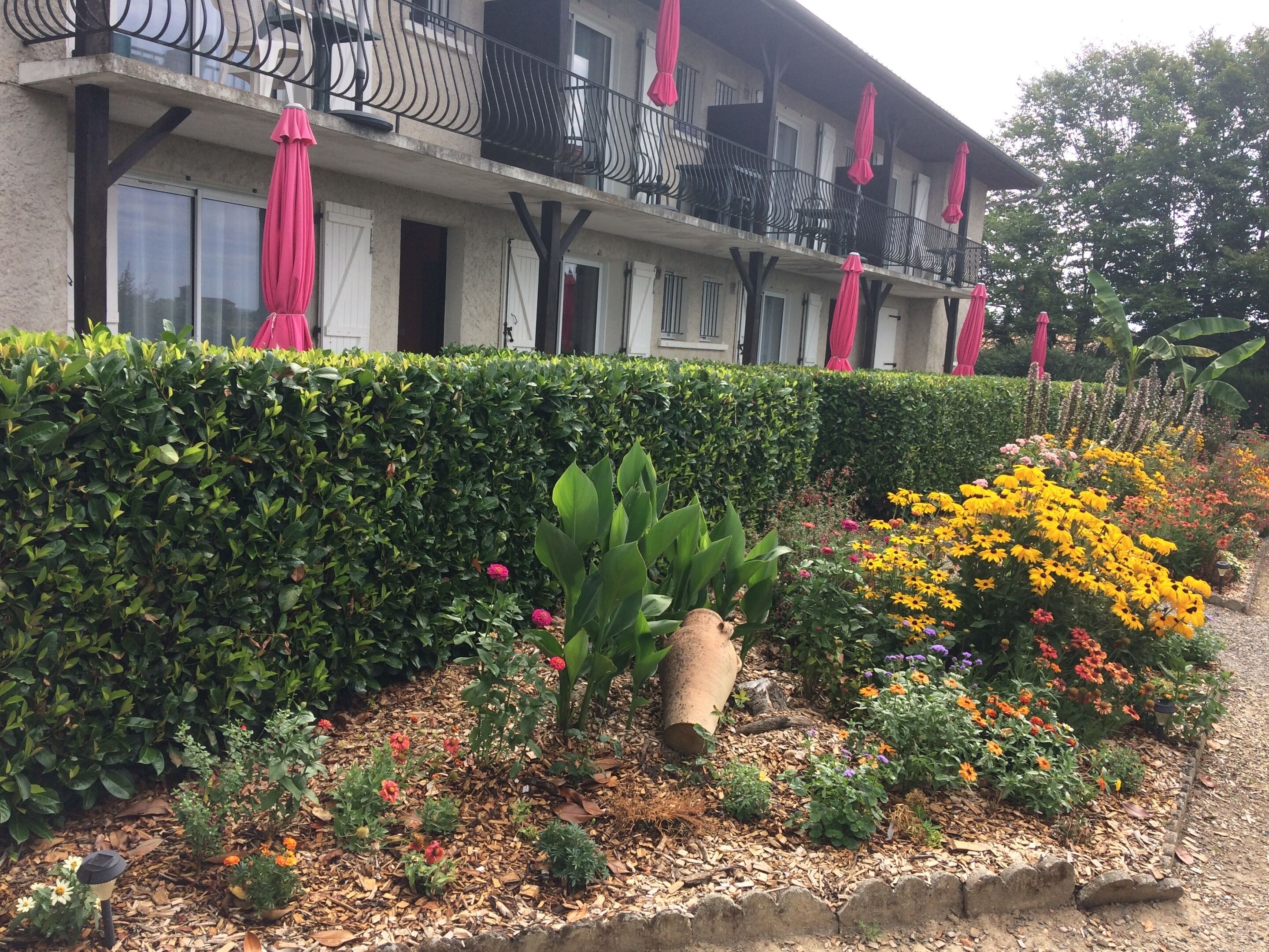 Een hotel met een balkon, een tuin vol planten en bloemen, en een haag.