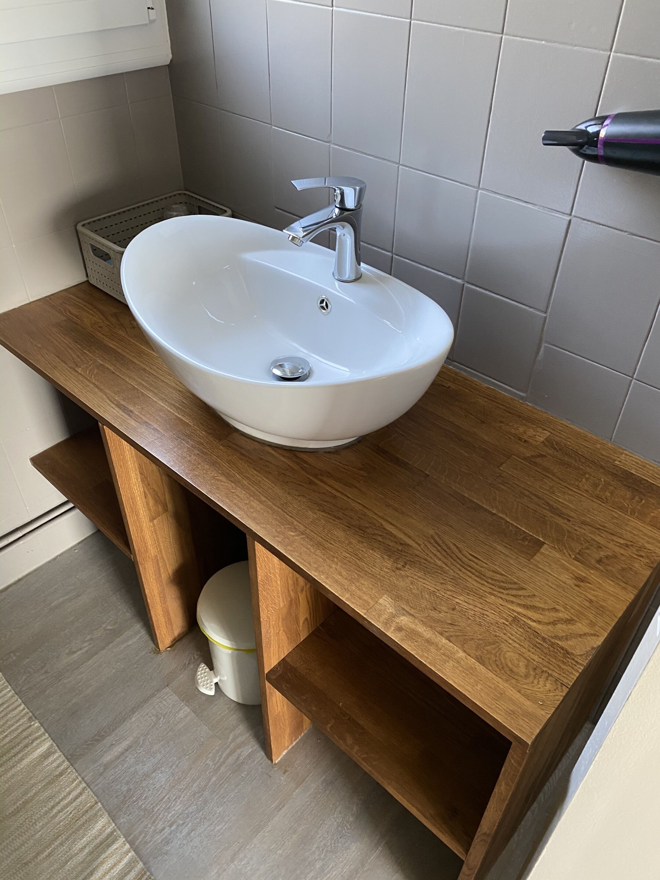 A bathroom with a white sink, wooden vanity, and grey tiled walls.
