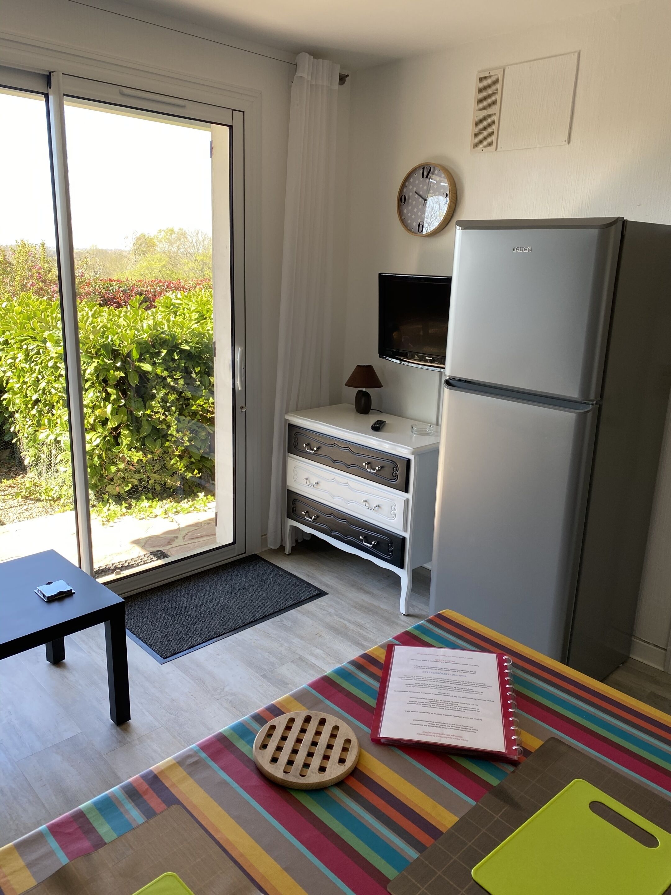 A compact kitchen with a refrigerator, a small table, a bench, and a view of greenery outside.