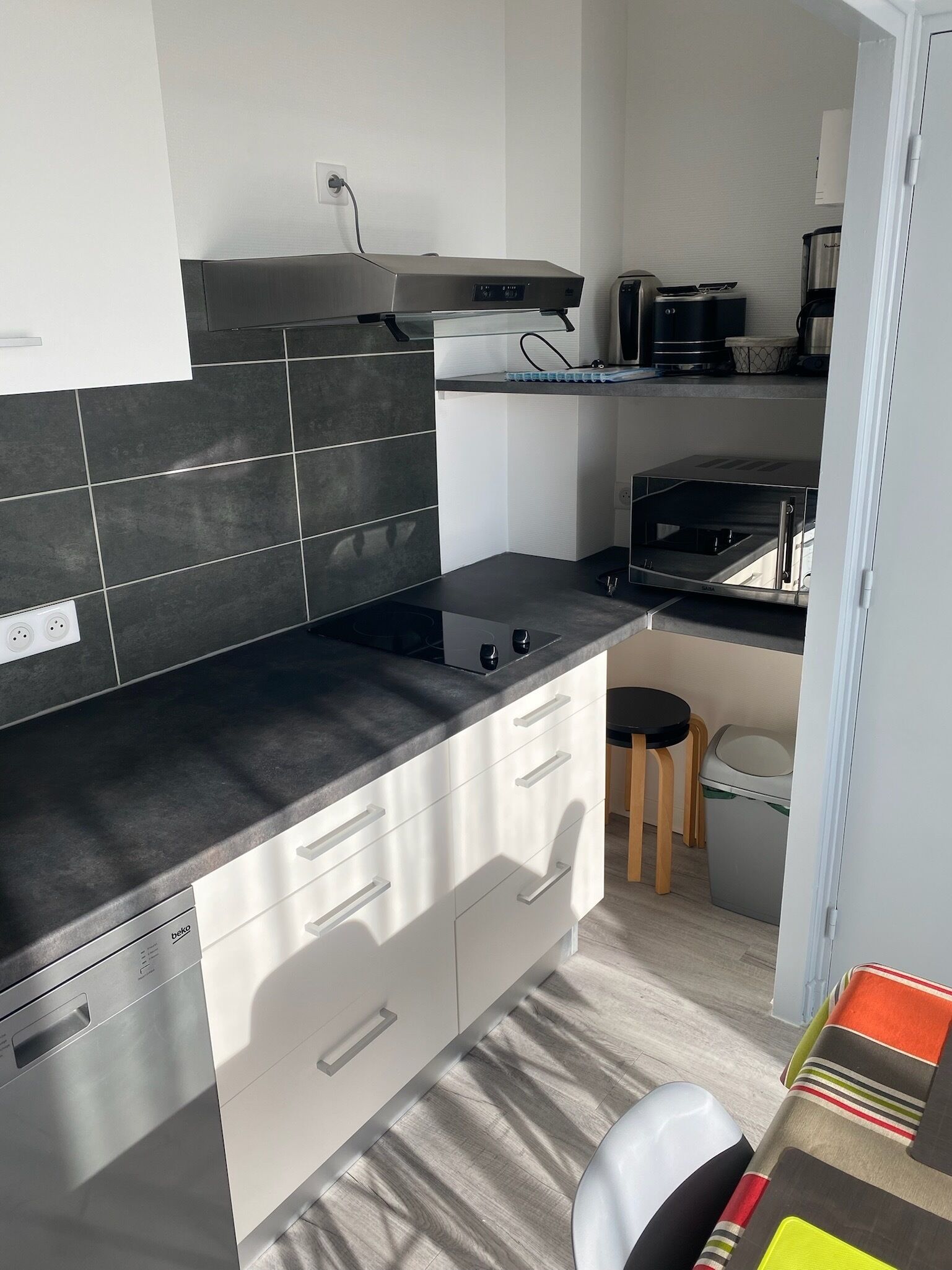 Una cucina compatta con un piano di lavoro nero, mobili bianchi, un forno integrato e un piccolo sgabello.