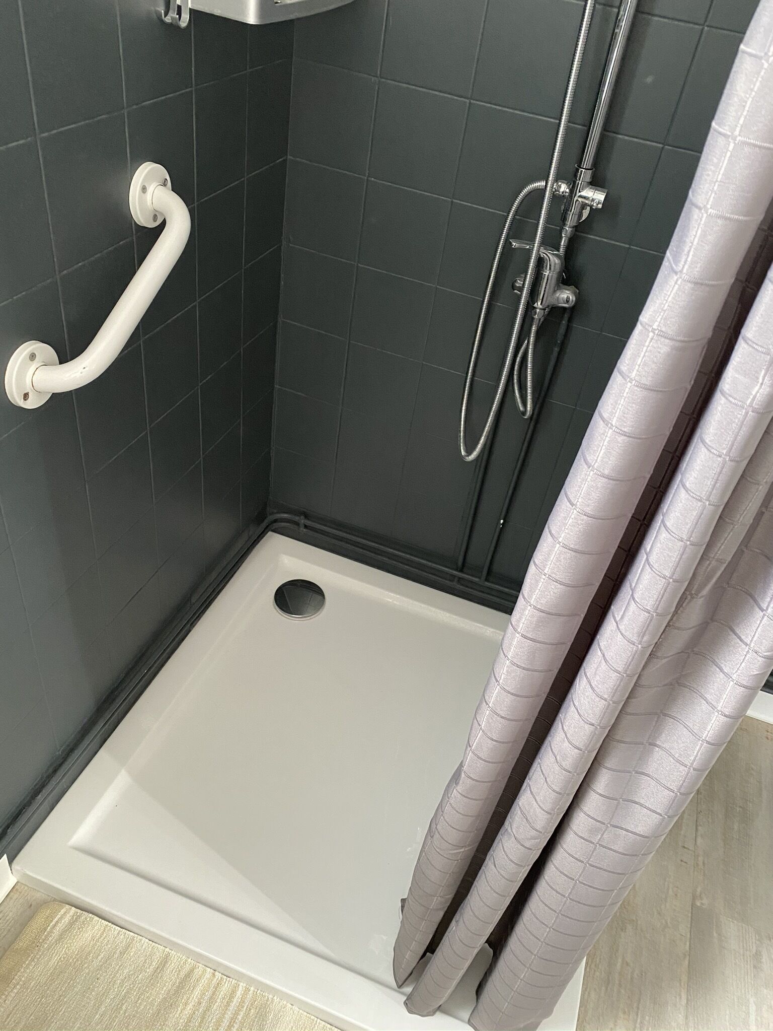 A small, enclosed shower area with a white grab bar, a showerhead, and a drain.