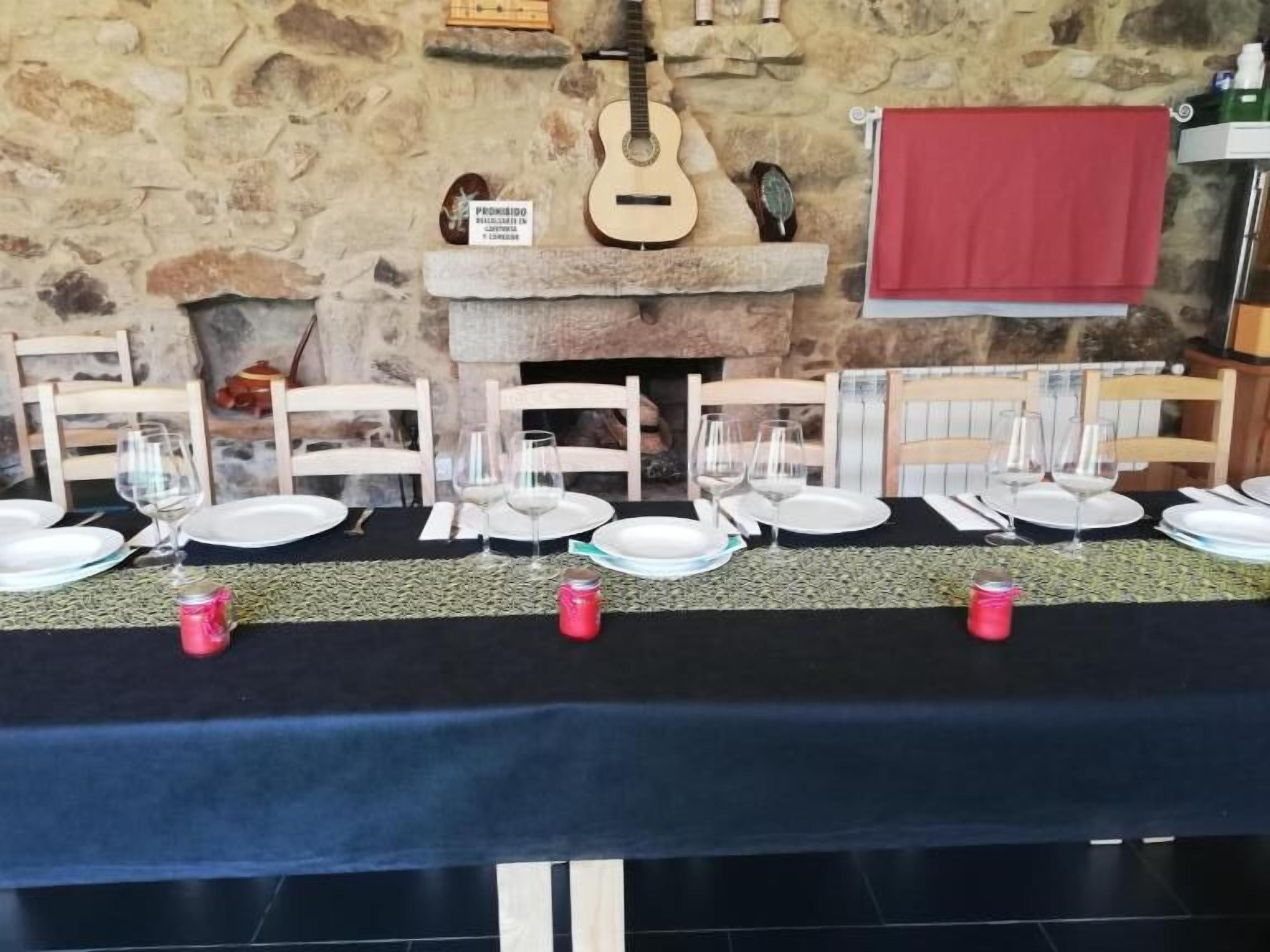 Taula de menjador preparada per a sis amb plats i gots blancs, una guitarra penjada a la paret i un fons de pared de pedra.
