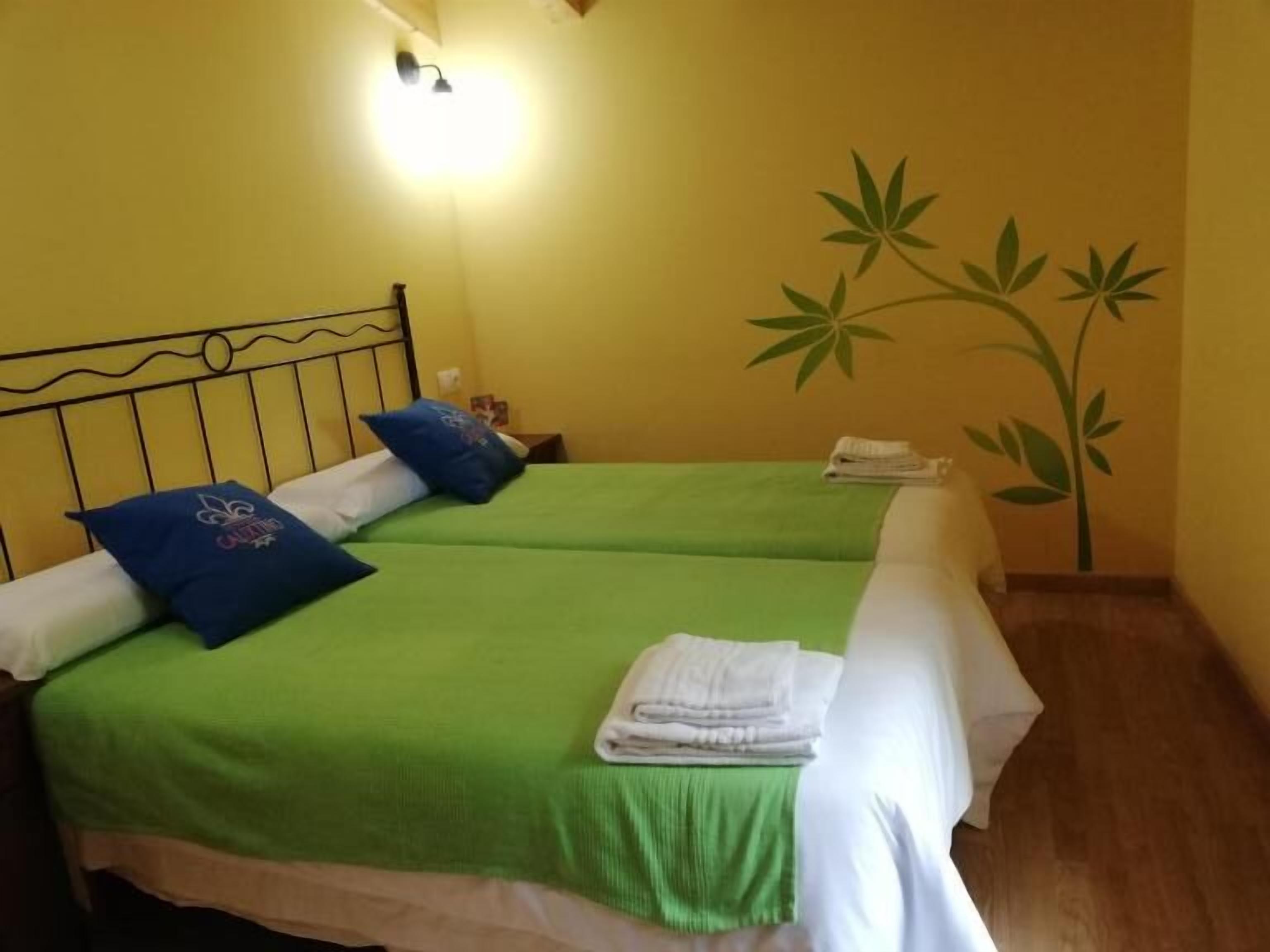 Een kamer met twee bedden, elk met groene beddengoed en blauwe kussens, een wandlamp en een muurschildering van een plant.