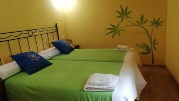 Een kamer met twee bedden, elk met groene beddengoed en blauwe kussens, een wandlamp en een muurschildering van een plant.